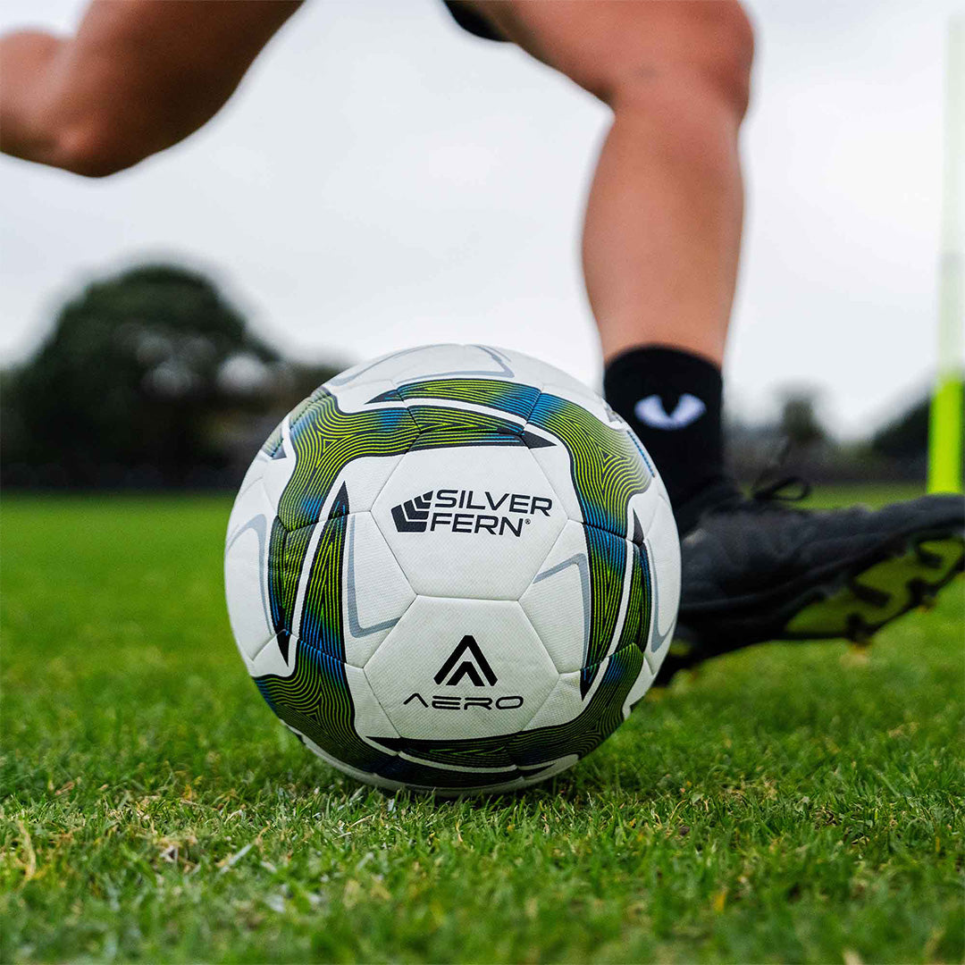 A close-up of an Aero size 5 soccer match ball with green and blue patterns, featuring the Silver Fern logo, placed on a grass field as a player in black soccer boots prepares to kick it.