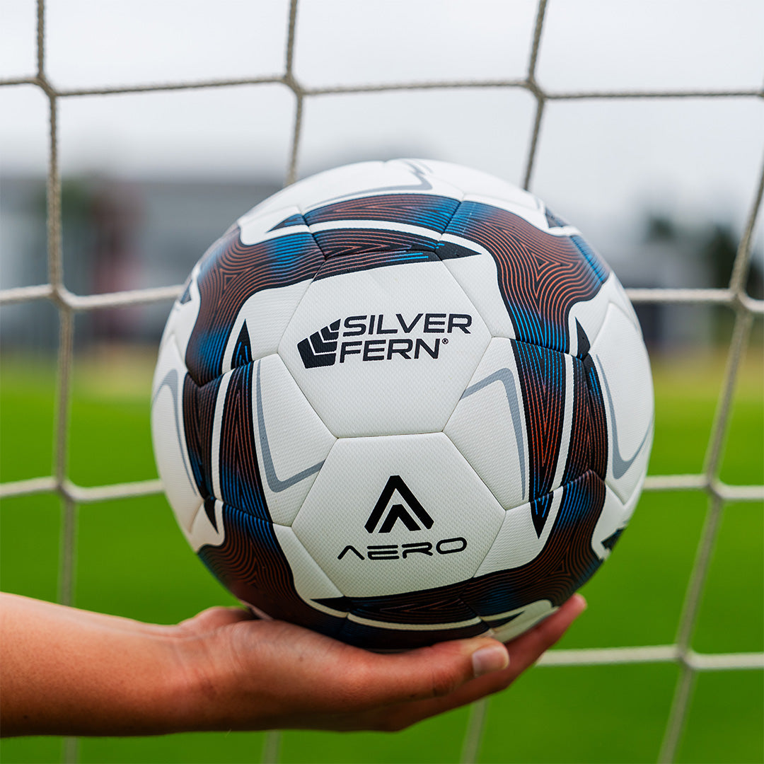 A close-up of an Aero size 4 soccer match ball being held by a player in front of a soccer goal.